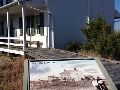 White two-story house with green shutters and an interpretive sign reading "Keep the Light Lit"