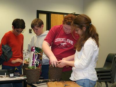 Four children repotting plants indoors; red shirt reads "CHURCHILL DOWNS".