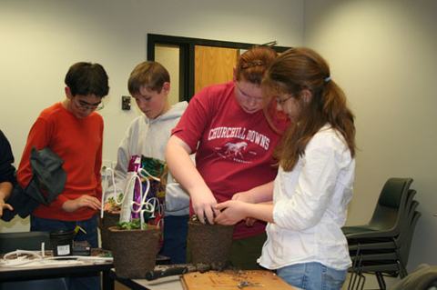 Four children repotting plants indoors; red shirt reads "CHURCHILL DOWNS".