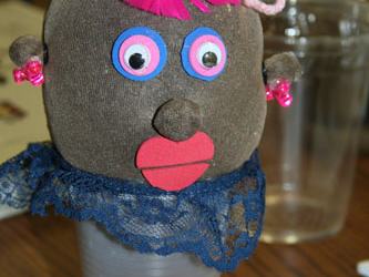 Sock puppet head with googly eyes, pink yarn hair and red lips atop a clear plastic cup