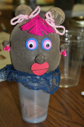 Sock puppet head with googly eyes, pink yarn hair and red lips atop a clear plastic cup