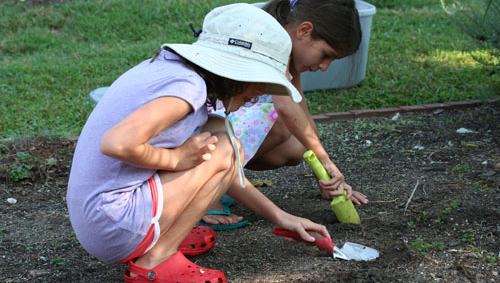 Two girls crouched digging soil with hand trowels beside a bucket