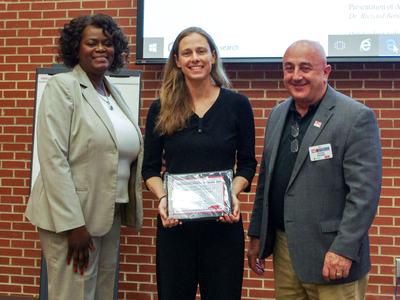 Woman poses in between two adults holding award in front of red brick background.