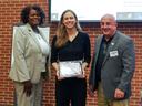 Woman poses in between two adults holding award in front of red brick background.