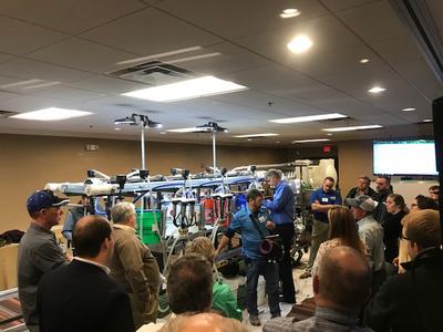 Group of people gathered around milking equipment during a demonstration in a conference room