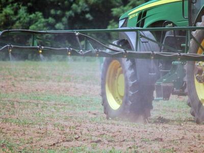 Green tractor with boom spraying mist over a bare field, rear wheels visible