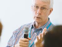 Older man with white hair and glasses holding a microphone while speaking at a meeting