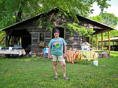 Woman standing in front of wooden barn beside tables; sign reads "Local Honey FOR SALE".