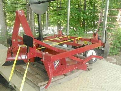 Red tractor-mounted tillage attachment secured with yellow straps on flat trailer
