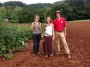Three people standing in a field holding an uprooted hemp plant; sign reads "Carmagnola Select"