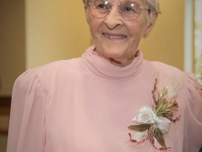 Elderly woman wearing glasses and a pink dress with floral corsage.