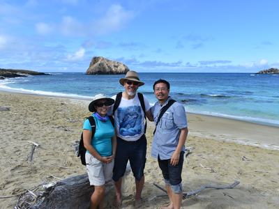 Three people standing on sandy beach by ocean and rock islets; center shirt reads "GSC"