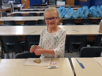 Child decorating a cookie with green icing at a cafeteria table, "Aim High" on shirt, "ICING" tube