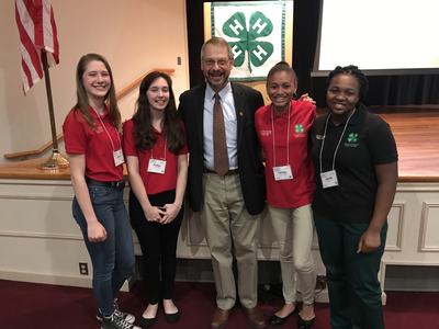 Five people posing: adult in suit with four youths wearing 4‑H shirts and nametags.