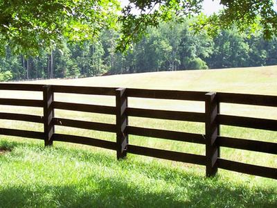 Dark wooden split-rail fence along sunlit grassy field with trees in background