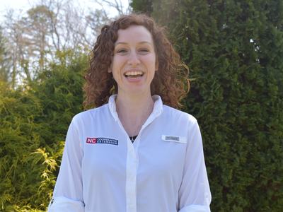 Smiling person with curly hair wearing white shirt labeled "NC Cooperative Extension" outdoors