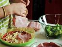 Hands placing ham slices onto a partially assembled pizza next to a bowl of broccoli