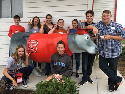 Eight teens posing around a red-and-gray cow statue with "NC" logo; one wears "CAROLINA" hoodie.