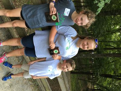 Three children standing in a wooded area holding 4‑H award plaques