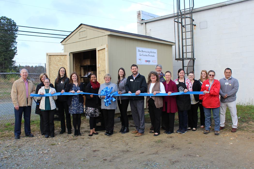 People holding blue ribbon for ribbon-cutting at shed labeled "The Borrowing Kitchen"
