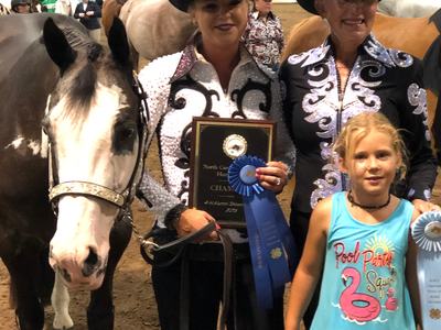 Two women in show jackets holding a plaque and blue ribbon beside a horse and child