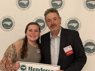 Two people holding a sign reading "Henderson" in front of North Carolina Association logos