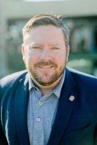 A man smiling with a coat and button up shirt on.