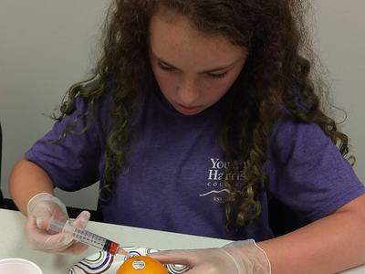 Child wearing gloves uses syringe to inject liquid into an orange on a paper plate.
