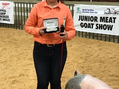 Girl holding award buckle and brush beside pig; banner reads "JUNIOR MEAT GOAT SHOW"