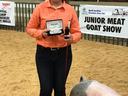 Girl holding award buckle and brush beside pig; banner reads "JUNIOR MEAT GOAT SHOW"