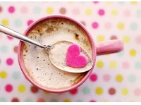 Pink mug of coffee with spoon holding a pink heart-shaped sugar on foam, polka-dot background