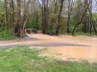 Flooded gravel path with muddy puddle leading to small concrete bridge amid trees