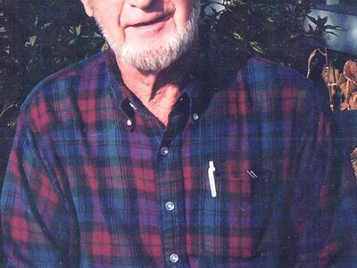 Older man with white beard seated outdoors wearing red-and-blue plaid shirt