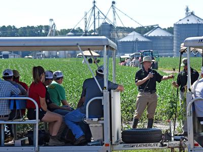 An NC State Extension specialist talking into a microphone in a soybean field while giving a tour to participants sitting in a tram.
