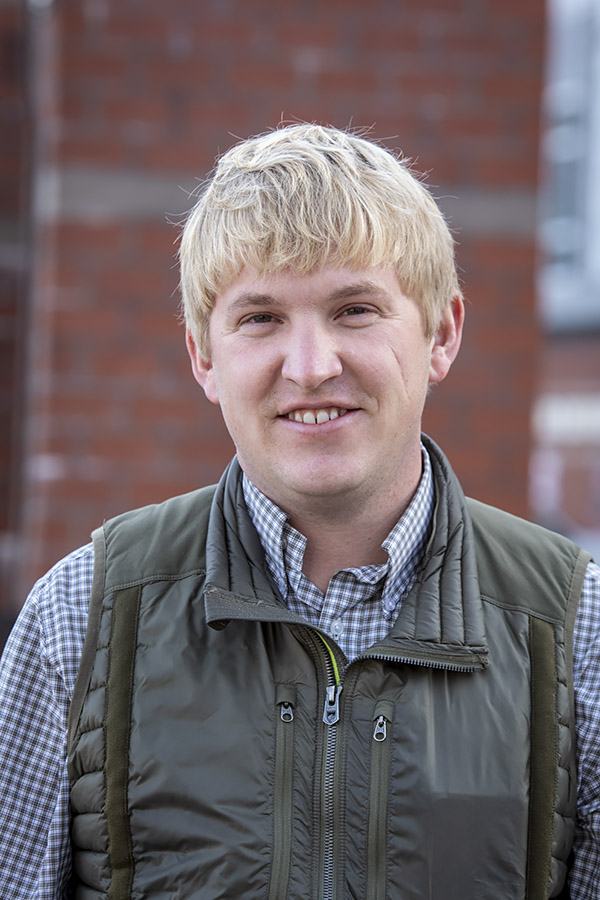 Man with blond hair in green quilted vest and checked shirt, brick wall background.