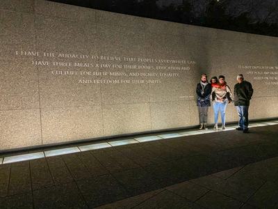 Four people stand by a wall engraved: "I have the audacity to believe that peoples everywhere can have three meals a day."