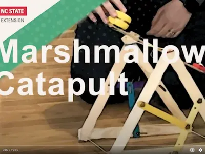 Person assembling a wooden marshmallow catapult; overlaid text "Marshmallow Catapult" and NC State Extension
