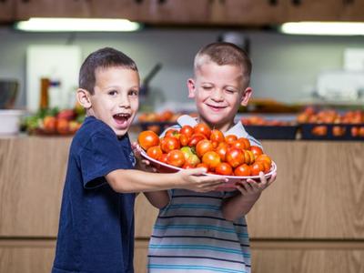 Two children holding a large bowl filled with tomatoes in a kitchen