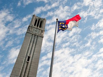 The North Carolina flag atop a flag pole beside the NC State University belltower, with a blue sky and white clouds above.
