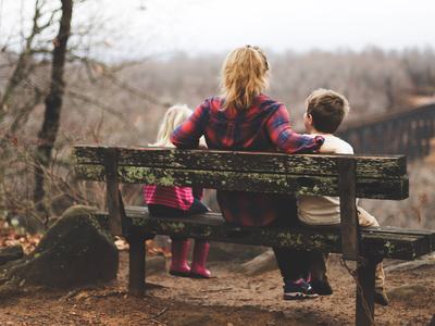 Adult and two children sitting on weathered bench overlooking forested valley and distant railroad trestle