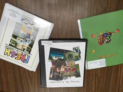 Three binders on a wooden table titled "FRESH", "Serving My Community", and "Cooking".