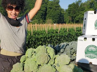 woman holding broccoli in the air and standing in front of a pile of broccoli heads