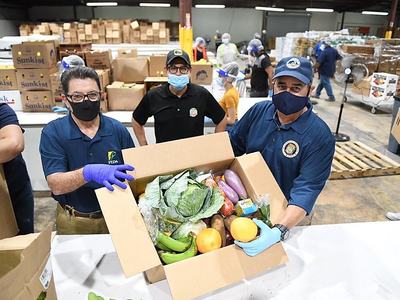 Volunteers in masks and gloves hold a box of assorted produce in a warehouse.