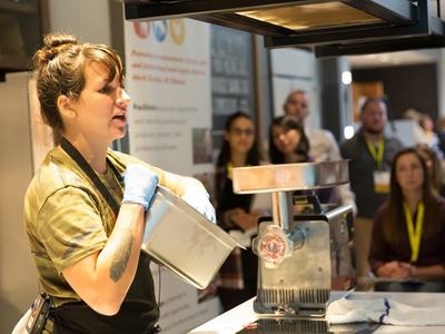 A woman demonstrates meat grinding equipment to an audience