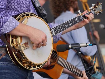 Musician strumming a banjo near a microphone, acoustic guitarist visible in background