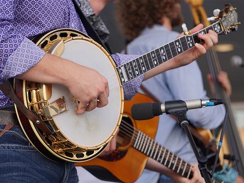 Musician strumming a banjo near a microphone, acoustic guitarist visible in background
