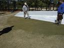 Three men inspecting turf beside a large white tarp on a grassy field