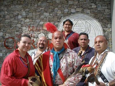 Six people in traditional regalia before a stone wall with partial text "MUSE" and "CHEROK"