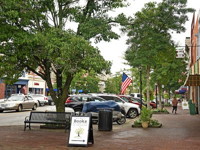 View of downtown city block. Trees line the brick sidewalks seperating the parking lot from the historic storefronts.