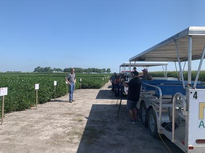 Man standing beside crop plots while camera crew films from a covered tram